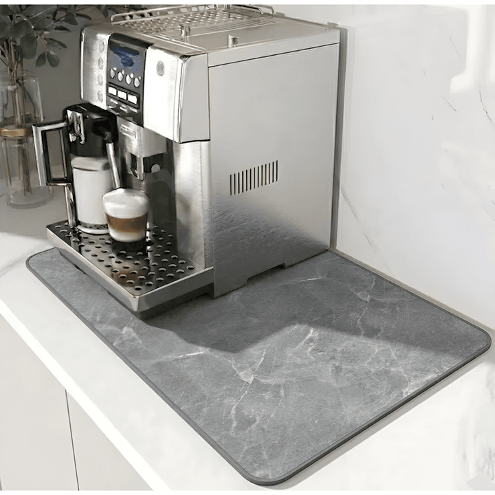 Graue Unterlage mit Marmormuster und dunklerem Rand unter einem Kaffeevollautomaten auf weißer Arbeitsplatte, links eine Pflanze in Glasvase. 12