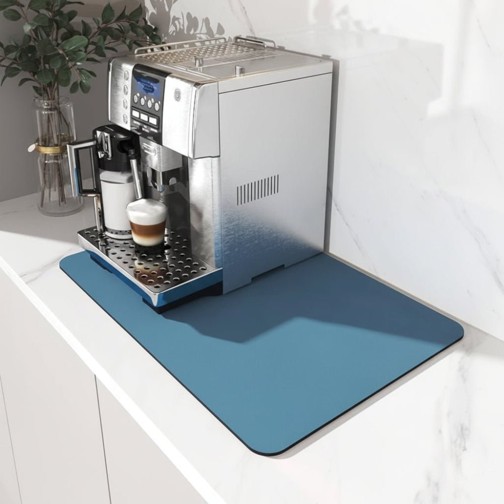 Blaue Unterlage mit abgerundeten Ecken unter einem Kaffeevollautomaten auf einer weißen Marmor-Arbeitsplatte, daneben eine Pflanze in einer Glasvase. 1