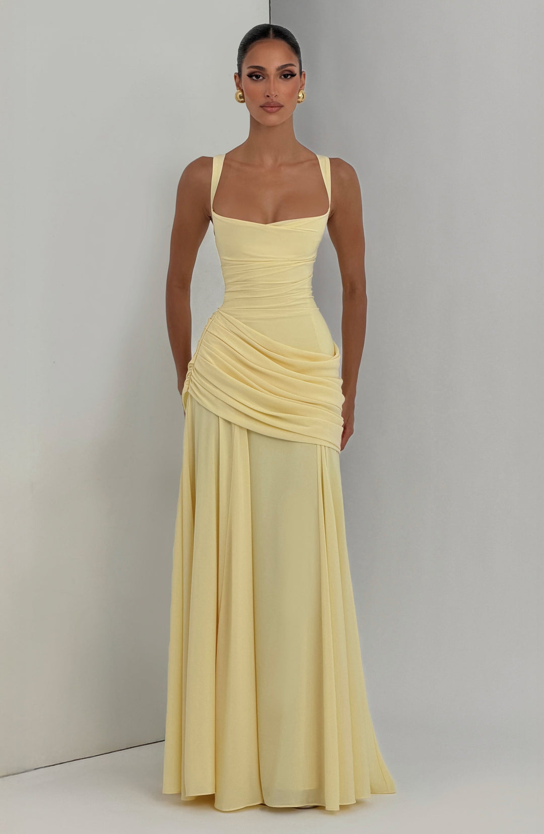 Model in hellgelbem Maxikleid mit goldenen Ohrringen, vor einer hellen Studiowand mit Ecke fotografiert. 6