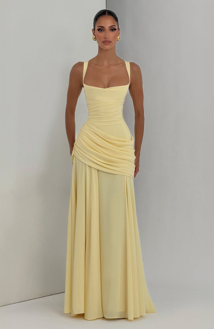 Model in hellgelbem Maxikleid mit goldenen Ohrringen, vor einer hellen Studiowand mit Ecke fotografiert. 6