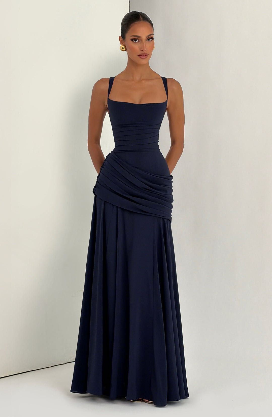 Model in marineblauem Maxikleid vor heller Studiowand mit Ecke, Hände in den Taschen, goldene Ohrringe. 13