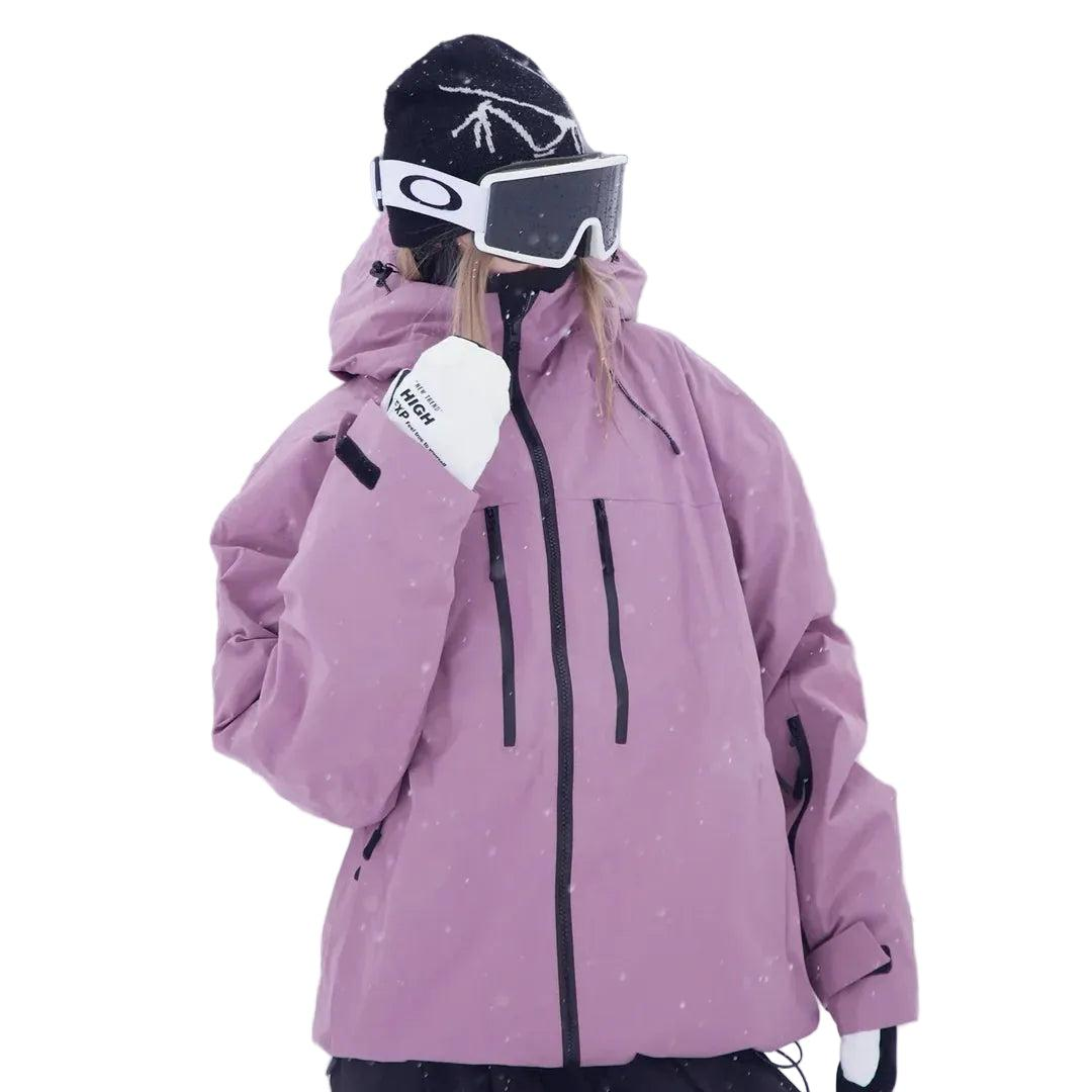 Frau in rosa-violetter Schneejacke mit Kapuze und Skibrille, die Hand vor dem Gesicht, Halbansicht mit Reißverschlusstaschen an der Brust. 2