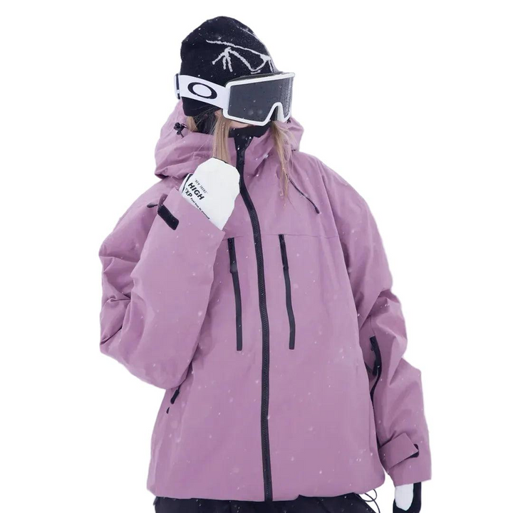 Frau in rosa-violetter Schneejacke mit Kapuze und Skibrille, die Hand vor dem Gesicht, Halbansicht mit Reißverschlusstaschen an der Brust. 2
