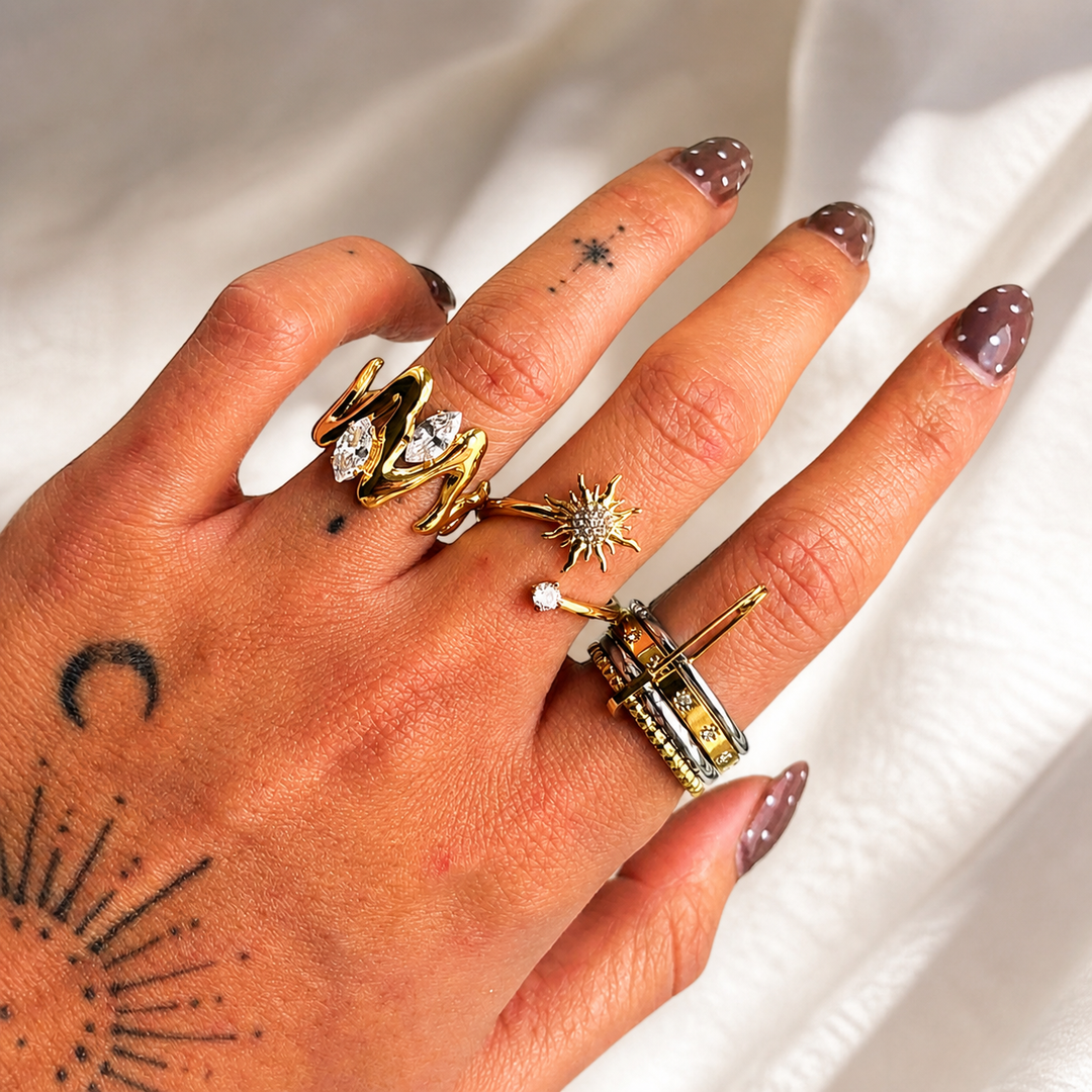 Hand mit tätowierten Motiven trägt den goldenen Wellenring am Zeigefinger, kombiniert mit weiteren Ringen, vor weißem Stoff. 3