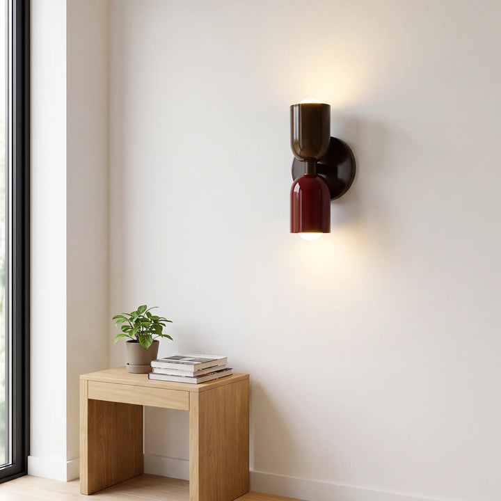 Wandlampe mit braunem oberem und weinrotem unterem Schirm an heller Wand, darunter ein Holzhocker mit Pflanze und Büchern am Fenster. 10