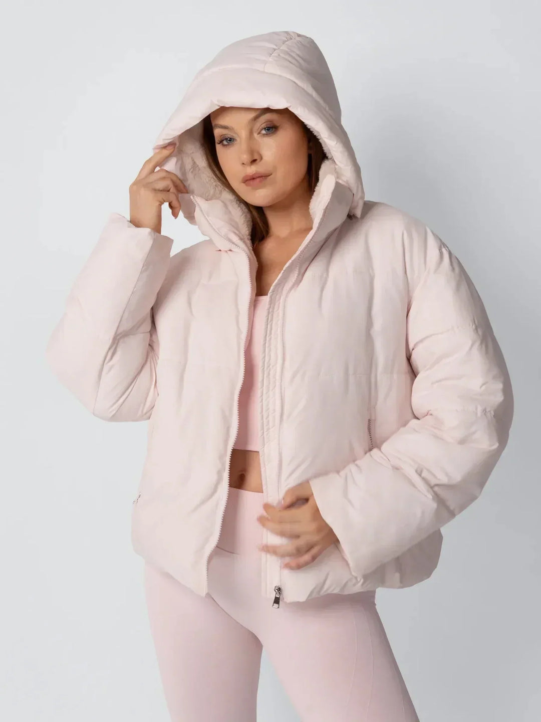 Model in rosafarbener Steppjacke mit hochgezogener Kapuze und Fleecefutter, eine Hand am Kapuzenrand 6