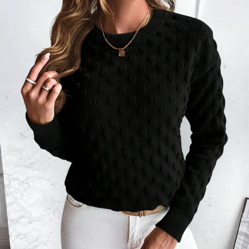 Frau in schwarzem Strickpullover mit Punktmuster, goldene Halskette, weiße Hose.