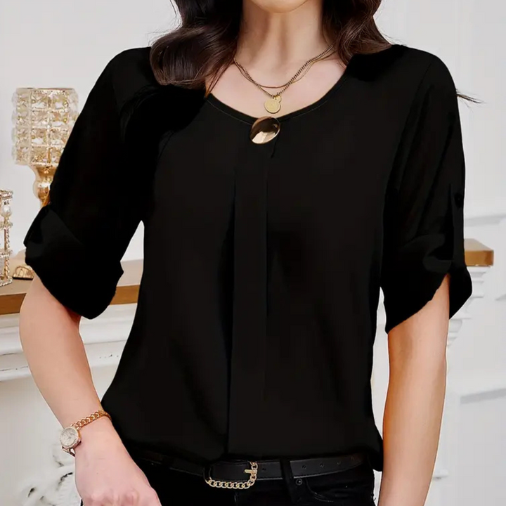 Frau in schwarzer Bluse mit Schmuck, stehend vor weißem Hintergrund, modisch elegant.