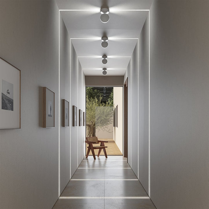 Vier weiße Deckenleuchten in einem Flur projizieren rechteckige Lichtlinien auf Wände, Decke und Boden, am Ende steht ein Stuhl. 1