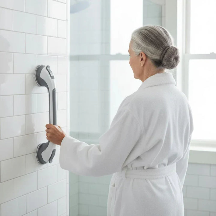 Frau im Bademantel hält sich in der gefliesten Dusche an einem senkrecht montierten Sauggriff fest, Rückansicht. 3