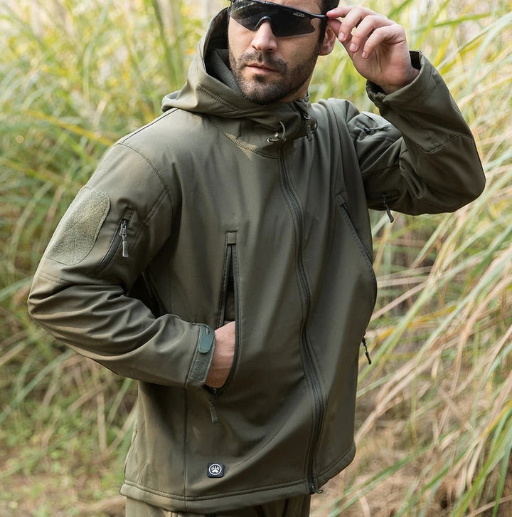 Mann in olivgrüner Jacke mit Sonnenbrille steht im hohen Gras und greift an die Brille, eine Hand in der Tasche. 11